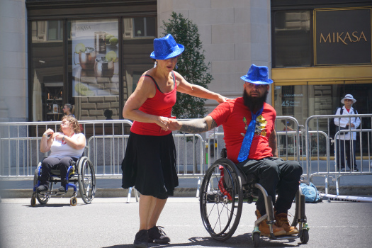 A woman and man are dancing wearing red tops, black bottoms, and blue hats. The man has a black beard and a wheelchair user.