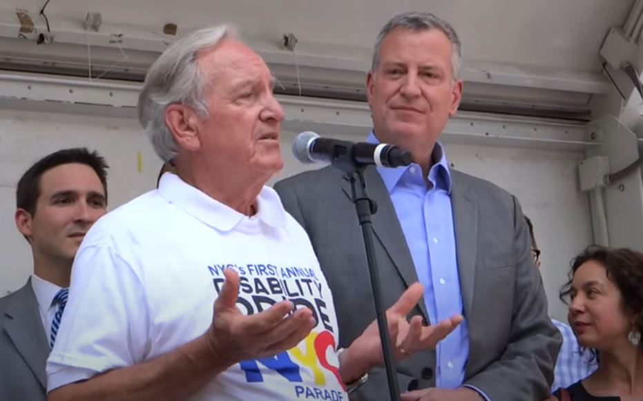 Two white men, one wearing a Disability Pride NYC t-shirt speaking into a Mic, the other wearing a shirt and jacket watching.