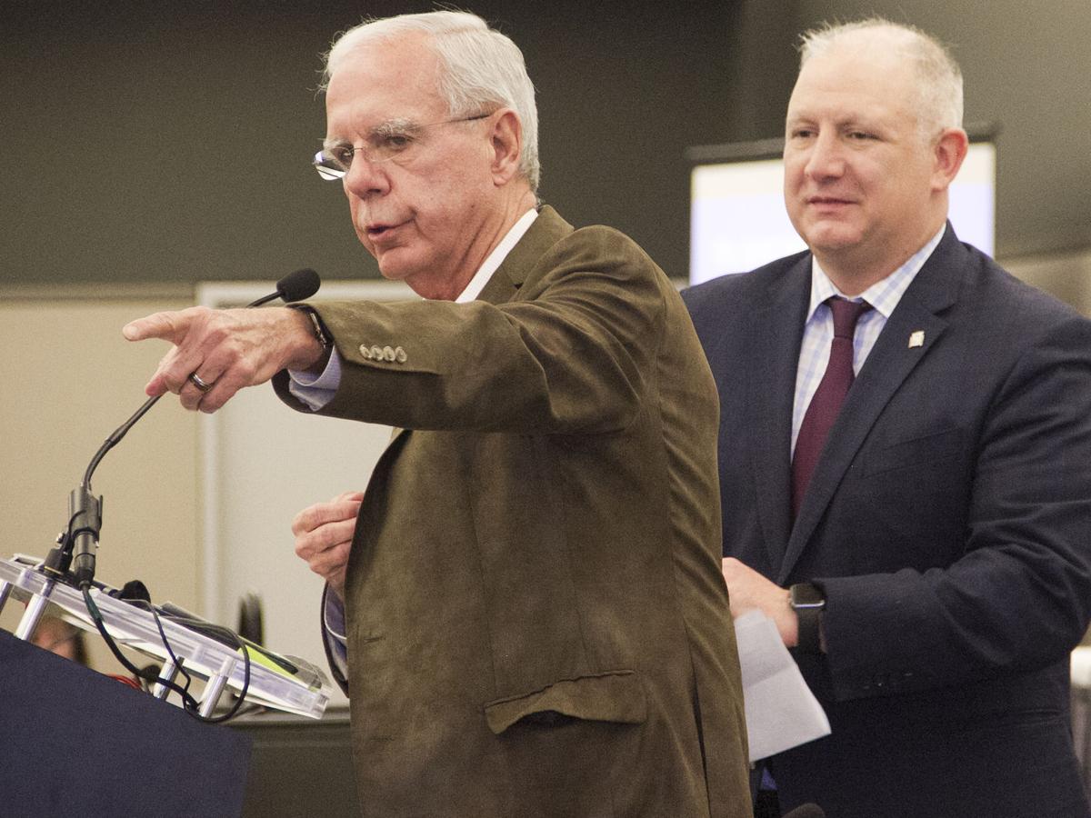 Two white men in suits, one speaking into a Mic pointing towards the camera the other stands behind him holding a paper.